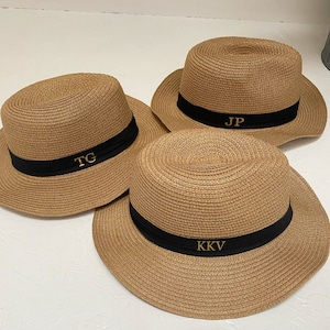 May include: Three beige straw hats with black bands. Each hat features gold embroidered initials: TC, JP, and KKV. The hats have a classic fedora shape with a wide brim, ideal for sun protection.