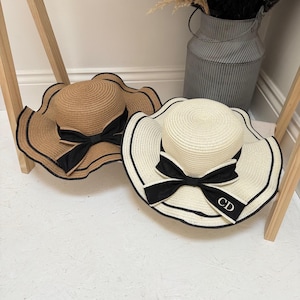 Può includere: Due cappelli di paglia da sole con fiocchi di nastro nero. Uno è marrone e l'altro è bianco. Il cappello bianco ha le iniziali "CD" ricamate sul nastro.