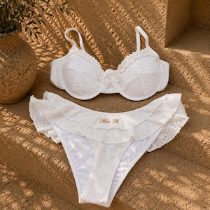 May include: A white two-piece bikini set made of eyelet fabric. The top features underwire cups with ruffled edges and thin straps. The bottom has a high-cut leg design with layered ruffles and the embroidered gold text "Mrs H" on the front.