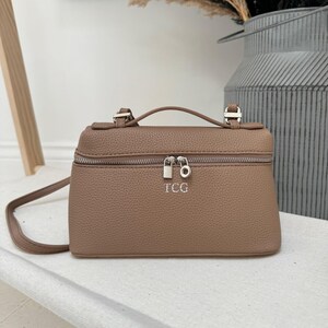 Puede incluir: Un bolso de mano satchel de piel sintética marrón con asa superior y bandolera. El bolso tiene cierre de cremallera y las iniciales TCG están grabadas en la parte delantera.