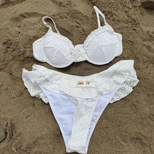 May include: White eyelet bikini set with ruffled edges. The top has underwire support and adjustable straps. The bottoms feature a high-cut design with a ruffle trim and the text "Mrs H". The bikini is displayed on a sandy surface.