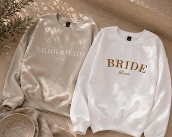 May include: A beige sweatshirt with the word BRIDESMAID and the name Amy embroidered in white, alongside a white sweatshirt with the word BRIDE and the name Laura embroidered in gold. Both feature crew necklines and long sleeves.