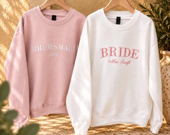 May include: A light pink sweatshirt with white embroidered text reading BRIDESMAID Lily and a white sweatshirt with pink embroidered text reading BRIDE Mrs Swift. Both crewneck garments feature long sleeves and are displayed on wooden hangers.