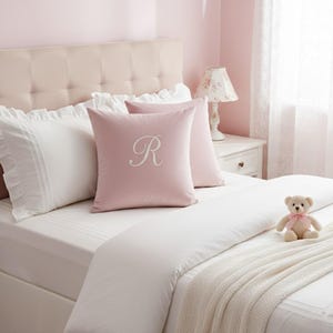May include: A bedroom scene with a bed featuring white bedding and pink pillows. One pink pillow has a white embroidered "R". A small teddy bear with a pink bow sits on the bed. A floral lamp and a white bedside table are visible.