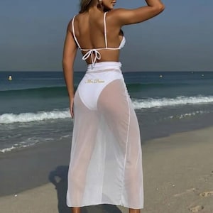 May include: A white bikini set with a matching sheer skirt. The bikini top has thin straps and ties at the back. The skirt is long and flowing, with the text "Mrs. Dunn" printed on the back of the bottoms. The outfit is worn on a beach.