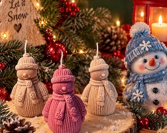 Pode incluir: Três velas em forma de boneco de neve, duas em bege claro e uma em rosa escuro, cada uma usando um gorro e cachecol de malha texturizada com detalhes de flocos de neve. As velas têm um pavio central e estão sobre uma fatia de madeira.