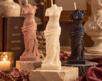 Op de afbeelding: Drie stompkaarsen in de vorm van het Venus van Milo-beeld in oudroze, wit en donkergrijs. Elke kaars heeft een lont, een gedrapeerd stofdetail op het onderlichaam en het woord VENUS in reliëf op de rechthoekige voet.