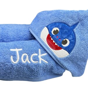 May include: A blue hooded towel featuring an embroidered shark face on the hood and the name Jack stitched in white thread on the front. The towel is made of a soft, textured terry cloth fabric with a matching blue trim along the edges.