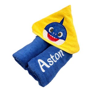 May include: A blue hooded towel with a yellow shark face embroidered on the hood. The towel is folded and the name "Aston" is embroidered on the towel.