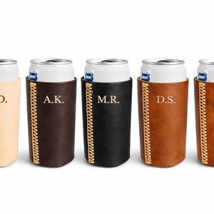 May include: Five personalized leather can coolers in various colors: tan, dark brown, black, medium brown, and light brown. Each cooler has initials stitched in gold thread and a matching gold-colored zipper.