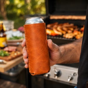 May include: A brown leather can cooler, held by a person, with a silver can inside. The cooler has visible stitching along the side. A grill with food is in the background, suggesting an outdoor gathering.