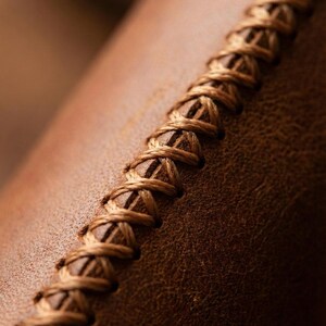 May include: Close-up of brown leather with a decorative, hand-stitched seam. The stitching is a lighter shade of brown, creating a textured contrast against the leather's surface. The image highlights the craftsmanship.