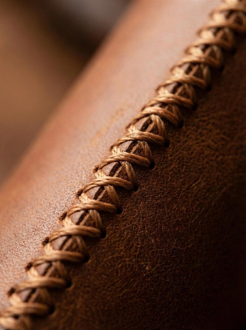 May include: Close-up of brown leather with a decorative, hand-stitched seam. The stitching is a lighter shade of brown, creating a textured contrast against the leather's surface. The image highlights the craftsmanship.