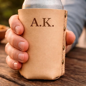 May include: A tan leather can cooler with the initials "A.K." engraved on the front. The cooler is held in a hand and has brass rivets along the side. A silver can is inside the cooler.