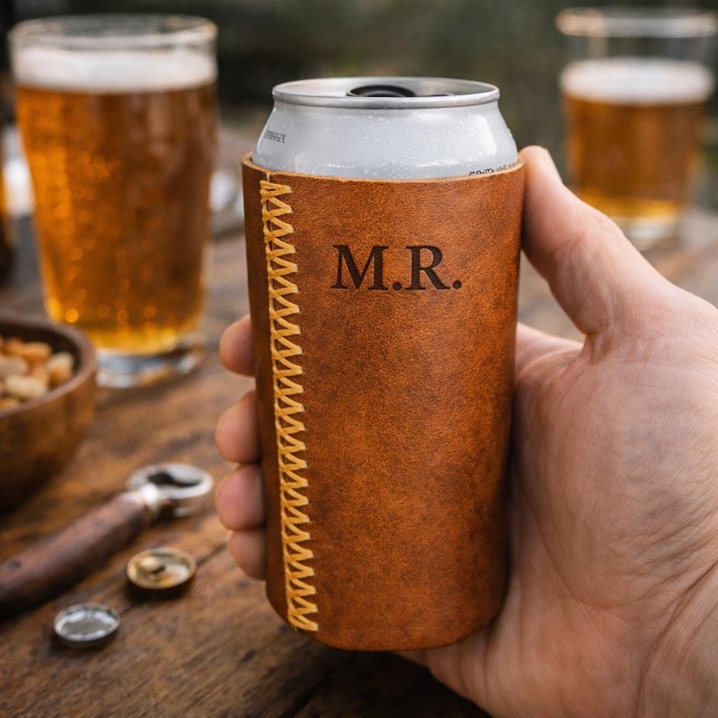 May include: A brown leather can cooler with yellow stitching and the initials "M.R." is held in a hand. The cooler is wrapped around a silver can. Two glasses of beer and a bowl of nuts are in the background.