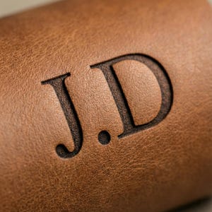 May include: Close-up of a brown leather item with the embossed initials "J.D" in a dark brown color. The leather has a textured surface, and the initials are centered.