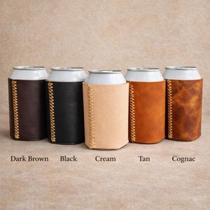May include: Five beverage can coolers in various colors: dark brown, black, cream, tan, and cognac. Each cooler is made of leather and features a stitched seam down the side. The colors are labeled below each cooler.