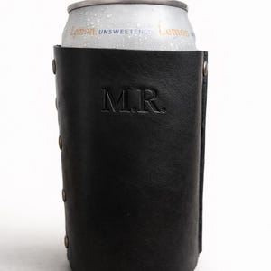 May include: A black leather can cooler with the initials "M.R." embossed on the front. The cooler holds a silver can of unsweetened lemon beverage. The cooler has rivets along the side.