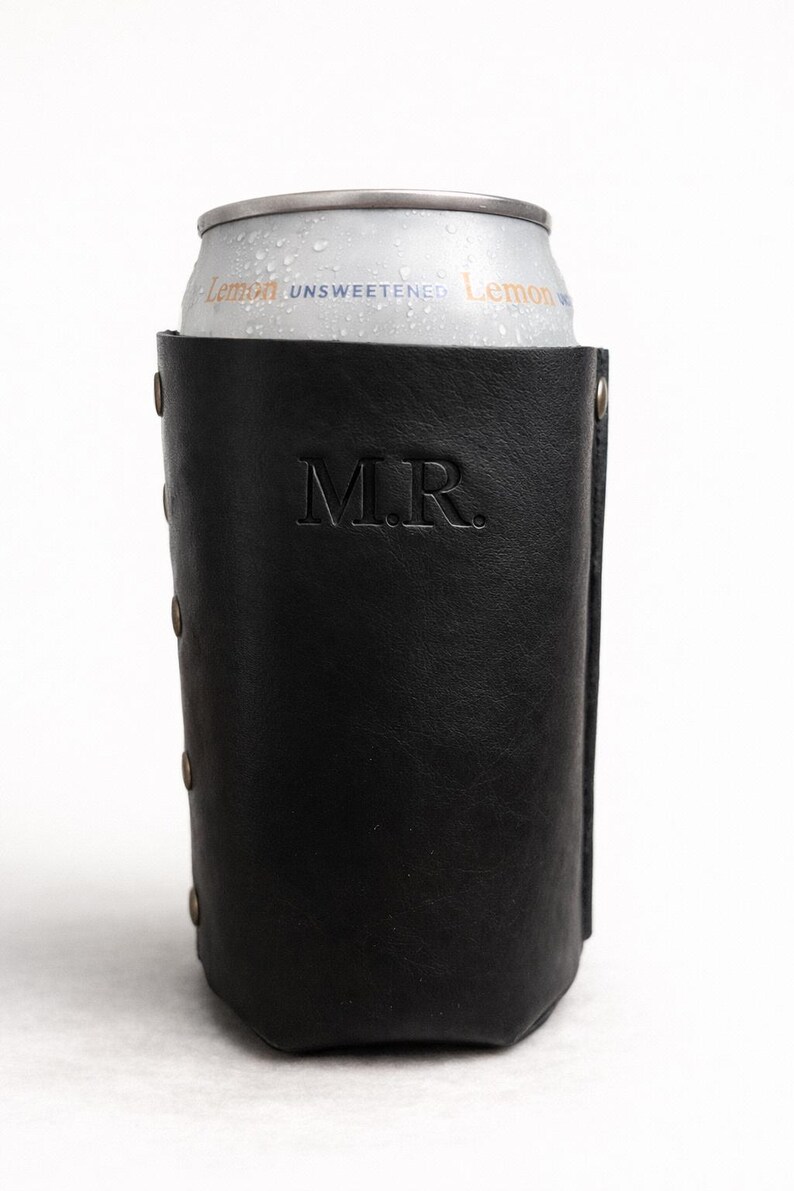 May include: A black leather can cooler with the initials "M.R." embossed on the front. The cooler holds a silver can of unsweetened lemon beverage. The cooler has rivets along the side.