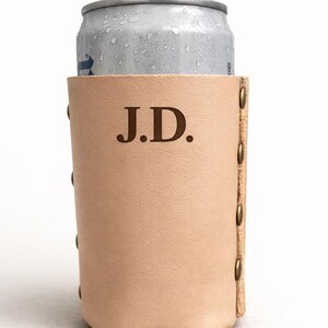 May include: A light tan leather can cooler with the initials "J.D." embossed on the front. The cooler is cylindrical, with brass rivets along the side. A silver can is partially visible inside the cooler.