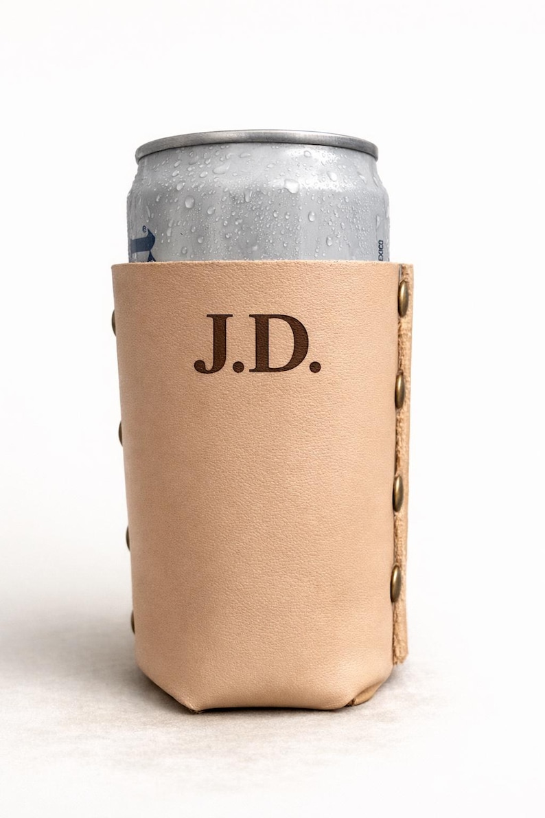 May include: A light tan leather can cooler with the initials "J.D." embossed on the front. The cooler is cylindrical, with brass rivets along the side. A silver can is partially visible inside the cooler.