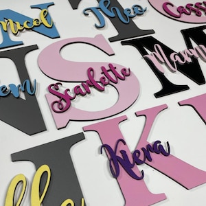 May include: A collection of wooden letters with names spelled out in various colours. The names include Theo, Cassie, Mamie, Scarlett, Kiena, and Em. The letters are painted in pink, purple, blue, yellow, black, and grey.