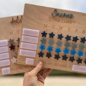 May include: Wooden star chart with the name "Jaxon's" at the top. The chart has spaces for stars and the days of the week. Includes blue, green, and black star-shaped tokens. A second chart reads "Add STAR" and "FOR STARS MY".