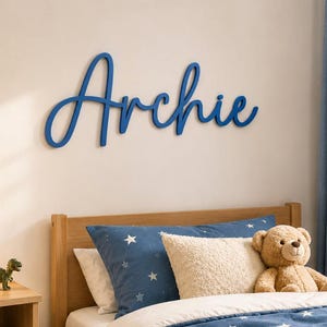 May include: A blue wall-mounted sign featuring the name Archie in a cursive script font. The letters are connected and have a smooth, matte finish. The sign is mounted horizontally on a plain wall above a bed with blue star-patterned bedding.