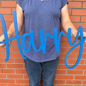 May include: A blue wooden sign spelling the name "Harry" in a cursive font. The sign is held in front of a person wearing a blue top and jeans, against a red brick wall background. The sign is a decorative item.