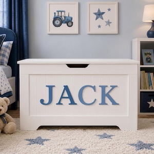May include: A white wooden toy chest featuring a rectangular shape with a horizontal cutout handle on the lid. The front panel has vertical slats and the name JACK displayed in large, bold, blue capital letters centred on the front surface.