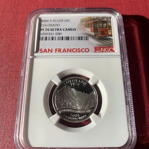 Peut inclure: Une pièce de monnaie de 25 cents en argent de 2006-S, classée PF 70 Ultra Cameo par NGC. La pièce représente le Capitole de l'État du Colorado et porte l'inscription "COLORADO 1876". La pièce est enfermée dans un support en plastique transparent avec une étiquette blanche qui indique "2006 S SILVER 25C COLORADO PF 70 ULTRA CAMEO 6299341-080 SAN FRANCISCO NGC".