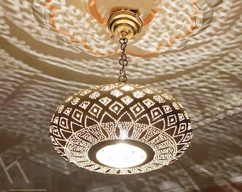 May include: A gold Moroccan pendant light with a geometric pattern casts intricate shadows on the ceiling. The light is hanging from a chain and has a white light bulb inside. The text "KeouCera -MOROCCAN HANDMADE- " is visible in the image.