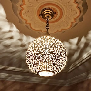 Puede incluir: Una lámpara colgante redonda de metal de color dorado con intrincados diseños recortados. La lámpara está colgada de una cadena y proyecta sombras en el techo.