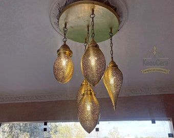 May include: A gold Moroccan chandelier with five lights hanging from a circular ceiling fixture. The lights are shaped like teardrops and have a decorative, patterned design. The chandelier is hanging in a room with a view of a swimming pool and a garden.