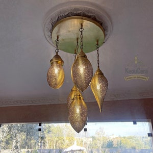 Puede incluir: Una lámpara de araña marroquí dorada con cinco luces que cuelgan de una fijación circular en el techo. Las luces tienen forma de lágrima y un diseño decorativo y estampado. La lámpara de araña está colgada en una habitación con vista a una piscina y un jardín.