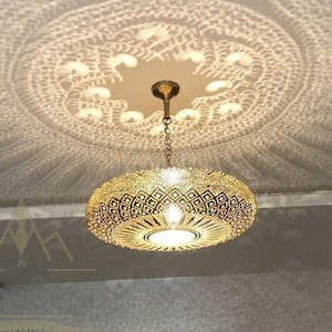Op de afbeelding: Een gouden plafondlamp in Marokkaanse stijl met ingewikkelde geometrische patronen. Het licht werpt een prachtig schaduwpatroon op het plafond.