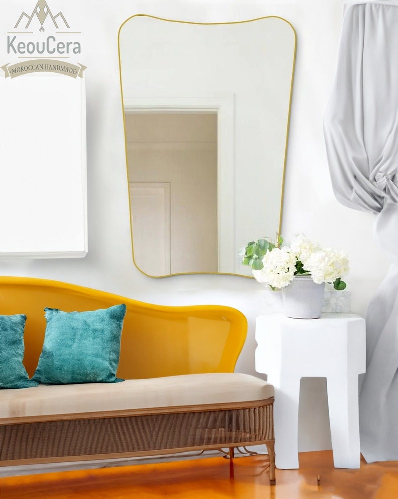 May include: A yellow curved sofa with a white cushion and two teal blue throw pillows. A white side table with a bouquet of white flowers sits next to the sofa. A large gold framed mirror hangs on the wall behind the sofa.