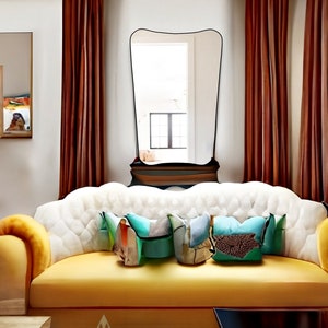 May include: A yellow sofa with white tufted upholstery and a curved back. The sofa has three colorful throw pillows and is positioned in front of a large, black-framed mirror. The mirror is set against a wall with brown curtains.