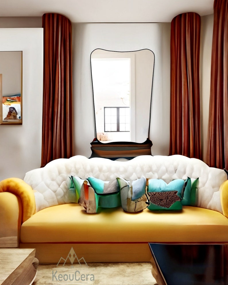 May include: A yellow sofa with white tufted upholstery and a curved back. The sofa has three colorful throw pillows and is positioned in front of a large, black-framed mirror. The mirror is set against a wall with brown curtains.