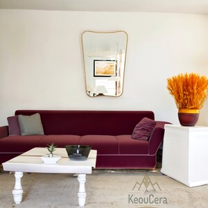 May include: A burgundy velvet sofa with gray and burgundy throw pillows sits in a living room with a white coffee table, a decorative vase with orange dried flowers, and a gold-framed mirror on the wall. The coffee table has a white ceramic bowl and a small potted plant.