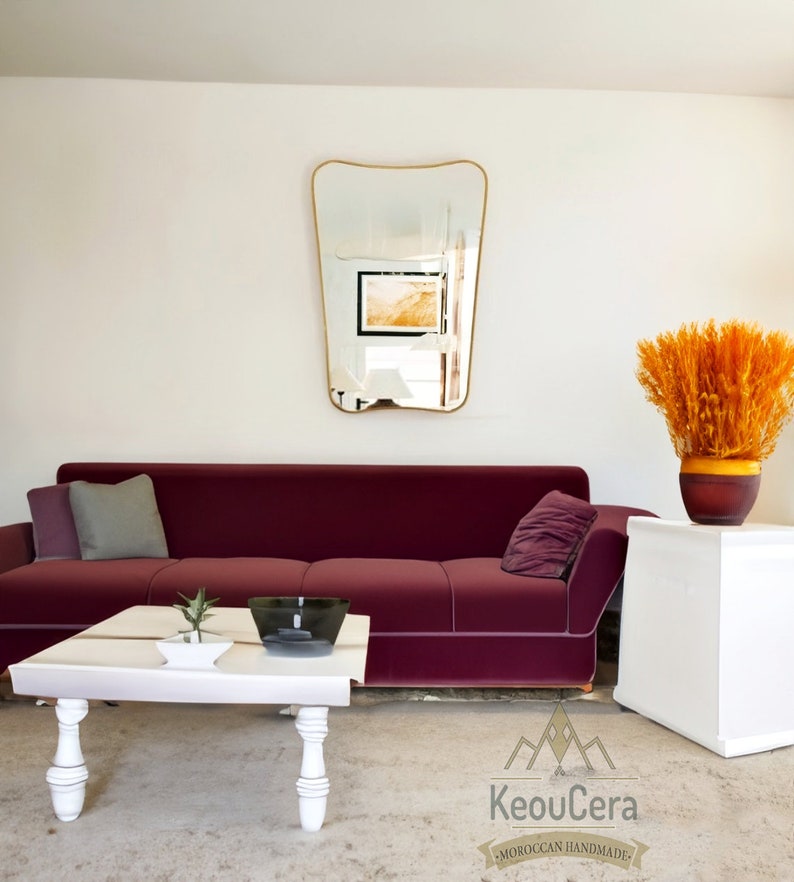 May include: A burgundy velvet sofa with gray and burgundy throw pillows sits in a living room with a white coffee table, a decorative vase with orange dried flowers, and a gold-framed mirror on the wall. The coffee table has a white ceramic bowl and a small potted plant.