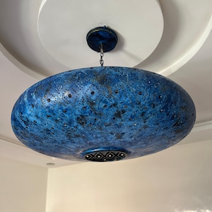 Può includere: Una lampada a sospensione ovale blu. L'apparecchio è sospeso al soffitto tramite una catena e presenta un design traforato con piccoli accenti scuri. La luce ha un aspetto strutturato e screziato e un motivo decorativo sul fondo.