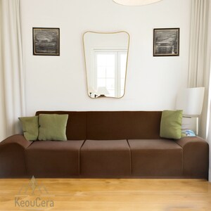 May include: A brown modular sofa with two green throw pillows. The sofa is in a modern living room with a white wall, a gold mirror, and two black and white framed pictures.