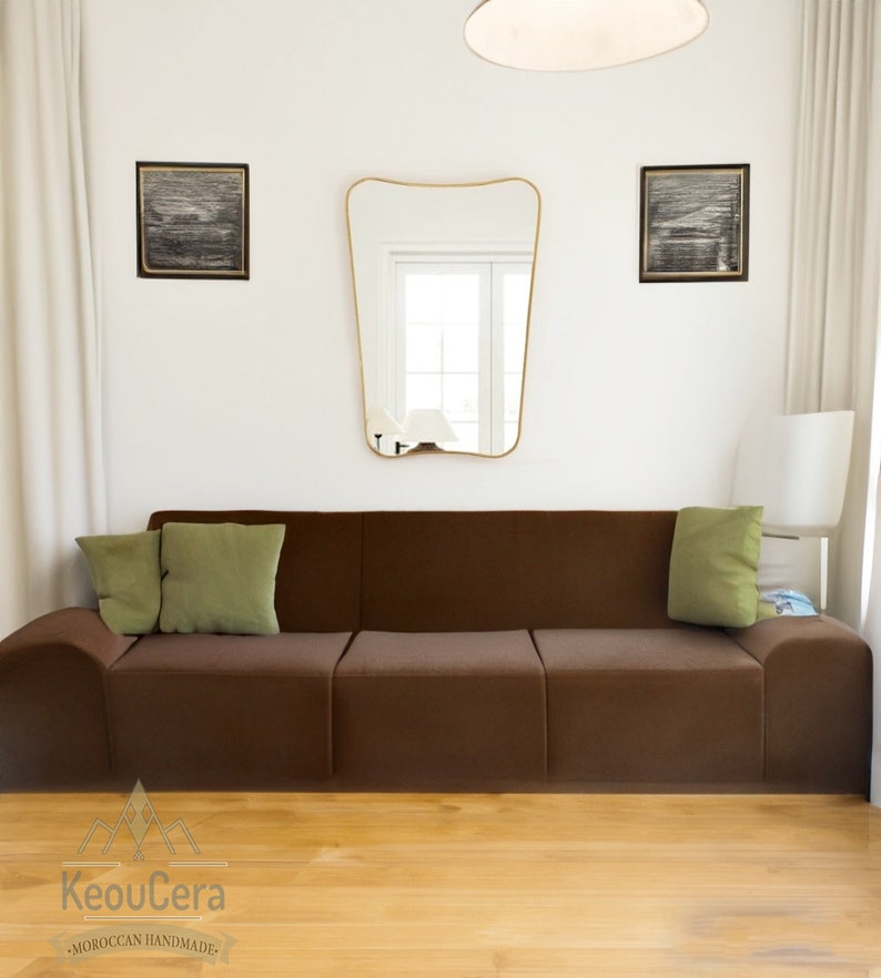 May include: A brown modular sofa with two green throw pillows. The sofa is in a modern living room with a white wall, a gold mirror, and two black and white framed pictures.