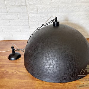May include: Black metal pendant light with a hammered finish and a chain hanging from the ceiling. The light is hanging over a wooden surface.