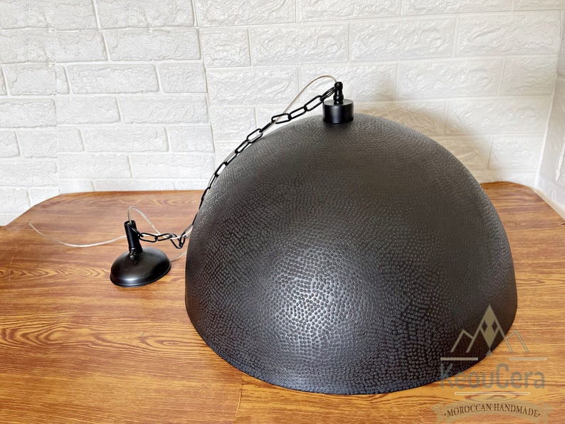 May include: Black metal pendant light with a hammered finish and a chain hanging from the ceiling. The light is hanging over a wooden surface.