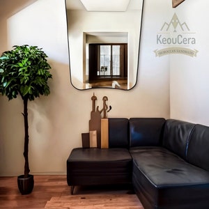 May include: A black leather sectional sofa with a curved mirror reflecting a room with a window. The mirror has a black frame and is mounted on a white wall. The sofa is in a corner of the room and there is a potted plant in the foreground.
