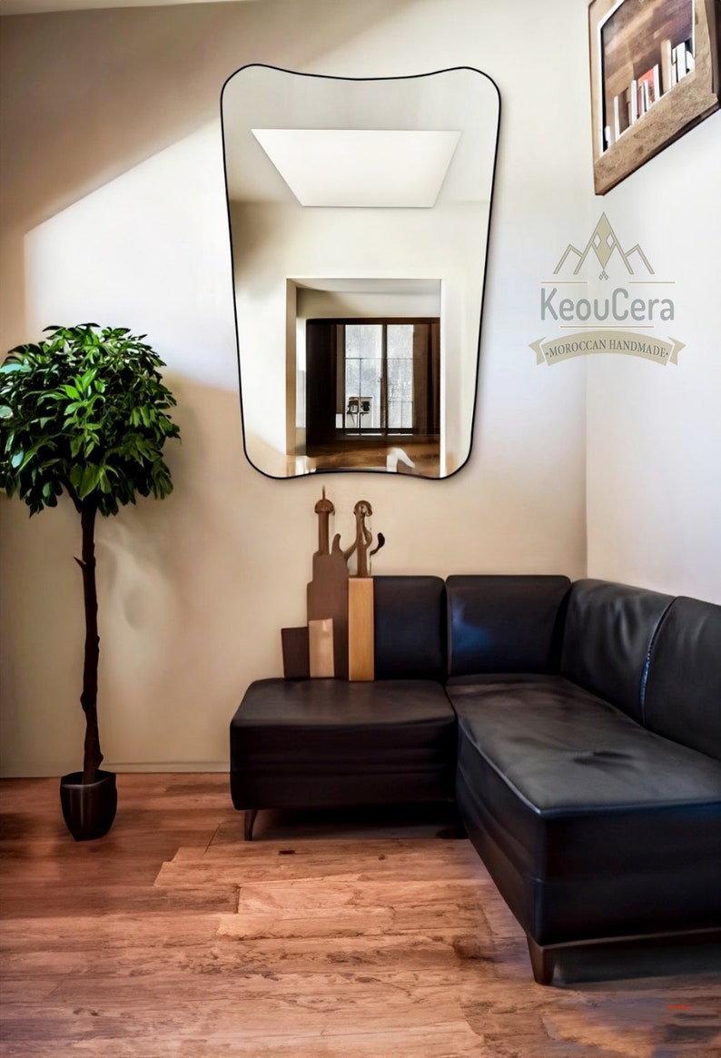 May include: A black leather sectional sofa with a curved mirror reflecting a room with a window. The mirror has a black frame and is mounted on a white wall. The sofa is in a corner of the room and there is a potted plant in the foreground.