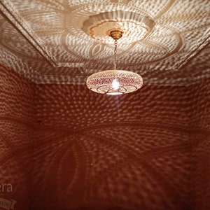 May include: A round, metal Moroccan pendant light fixture with a decorative, patterned design casts intricate shadows on the ceiling and walls of a room. The light is hanging from a chain and has a warm, white light bulb.