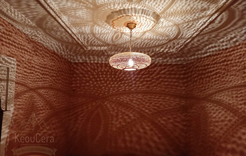 May include: A round, metal Moroccan pendant light fixture with a decorative, patterned design casts intricate shadows on the ceiling and walls of a room. The light is hanging from a chain and has a warm, white light bulb.